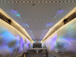A modern interior with an illuminated ceiling covered in small, recessed lights. The side walls display an artistic, wave-like texture with soft, pastel blue and purple hues. Large windows can be seen at the far end, partially reflecting the ambient light.