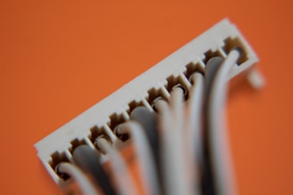 A close-up view of a white plastic connector attached to several grey wires against an orange background. The image focuses on the small pins inside the connector slots, showing fine details of its construction.