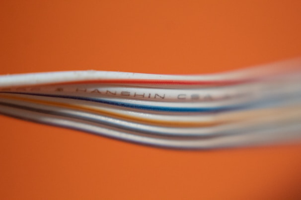 Several colored cables are stacked closely together against a bright orange background. The cables, including white, blue, and red, create a layered texture. Some text is visible on one of the cables, though it is partially blurred.