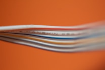 Several colored cables are stacked closely together against a bright orange background. The cables, including white, blue, and red, create a layered texture. Some text is visible on one of the cables, though it is partially blurred.