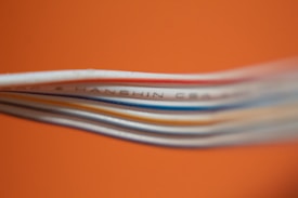 Several colored cables are stacked closely together against a bright orange background. The cables, including white, blue, and red, create a layered texture. Some text is visible on one of the cables, though it is partially blurred.