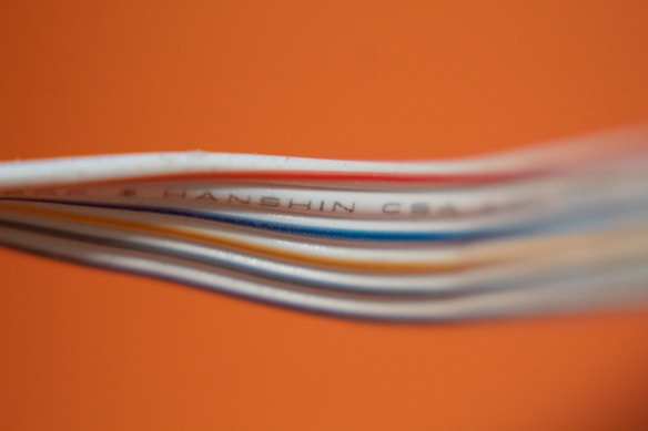 Several colored cables are stacked closely together against a bright orange background. The cables, including white, blue, and red, create a layered texture. Some text is visible on one of the cables, though it is partially blurred.