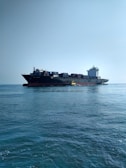 A large cargo ship carrying numerous containers is moving across a calm ocean. The ship is accompanied by a small yellow pilot boat near its side. The sky is clear, adding to the tranquility of the scene.