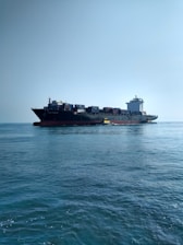 A large cargo ship carrying numerous containers is moving across a calm ocean. The ship is accompanied by a small yellow pilot boat near its side. The sky is clear, adding to the tranquility of the scene.