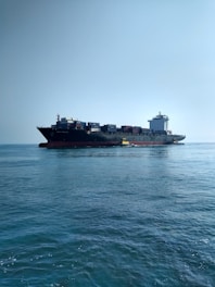 A large cargo ship carrying numerous containers is moving across a calm ocean. The ship is accompanied by a small yellow pilot boat near its side. The sky is clear, adding to the tranquility of the scene.