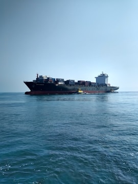 A large cargo ship carrying numerous containers is moving across a calm ocean. The ship is accompanied by a small yellow pilot boat near its side. The sky is clear, adding to the tranquility of the scene.
