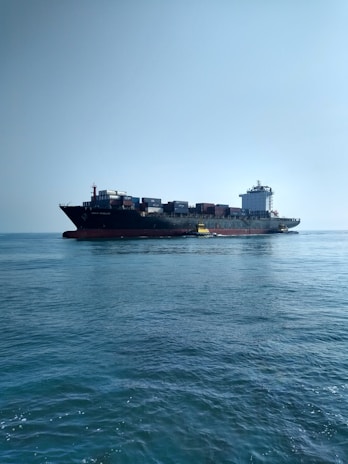 A large cargo ship carrying numerous containers is moving across a calm ocean. The ship is accompanied by a small yellow pilot boat near its side. The sky is clear, adding to the tranquility of the scene.