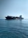A large cargo ship carrying numerous containers is moving across a calm ocean. The ship is accompanied by a small yellow pilot boat near its side. The sky is clear, adding to the tranquility of the scene.