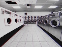 A laundromat interior is visible, featuring rows of industrial washing machines and dryers along two walls. The machines on the left are white, while the ones on the right are metallic. The floor consists of light gray tiles, and the ceiling has evenly spaced fluorescent lights. The overall environment is clean and orderly, with no people present.