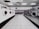 A laundromat interior is visible, featuring rows of industrial washing machines and dryers along two walls. The machines on the left are white, while the ones on the right are metallic. The floor consists of light gray tiles, and the ceiling has evenly spaced fluorescent lights. The overall environment is clean and orderly, with no people present.