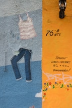 A colorful mural on a textured wall featuring a painted striped tank top and jeans. The right section of the wall is painted in orange with handwritten text in Spanish indicating opening hours and a welcoming message. There are decorative flower patterns and a black lantern is mounted above.