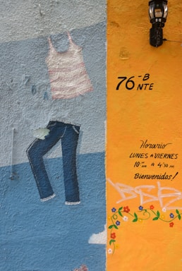 A colorful mural on a textured wall featuring a painted striped tank top and jeans. The right section of the wall is painted in orange with handwritten text in Spanish indicating opening hours and a welcoming message. There are decorative flower patterns and a black lantern is mounted above.