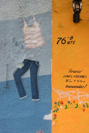A colorful mural on a textured wall featuring a painted striped tank top and jeans. The right section of the wall is painted in orange with handwritten text in Spanish indicating opening hours and a welcoming message. There are decorative flower patterns and a black lantern is mounted above.