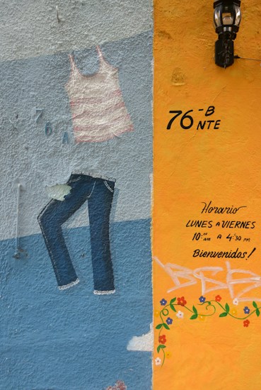 A colorful mural on a textured wall featuring a painted striped tank top and jeans. The right section of the wall is painted in orange with handwritten text in Spanish indicating opening hours and a welcoming message. There are decorative flower patterns and a black lantern is mounted above.