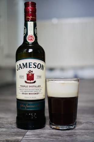 A bottle of Jameson Triple Distilled Irish Whiskey is placed on a surface next to a glass filled with a dark liquid topped with a layer of cream, resembling an Irish coffee.
