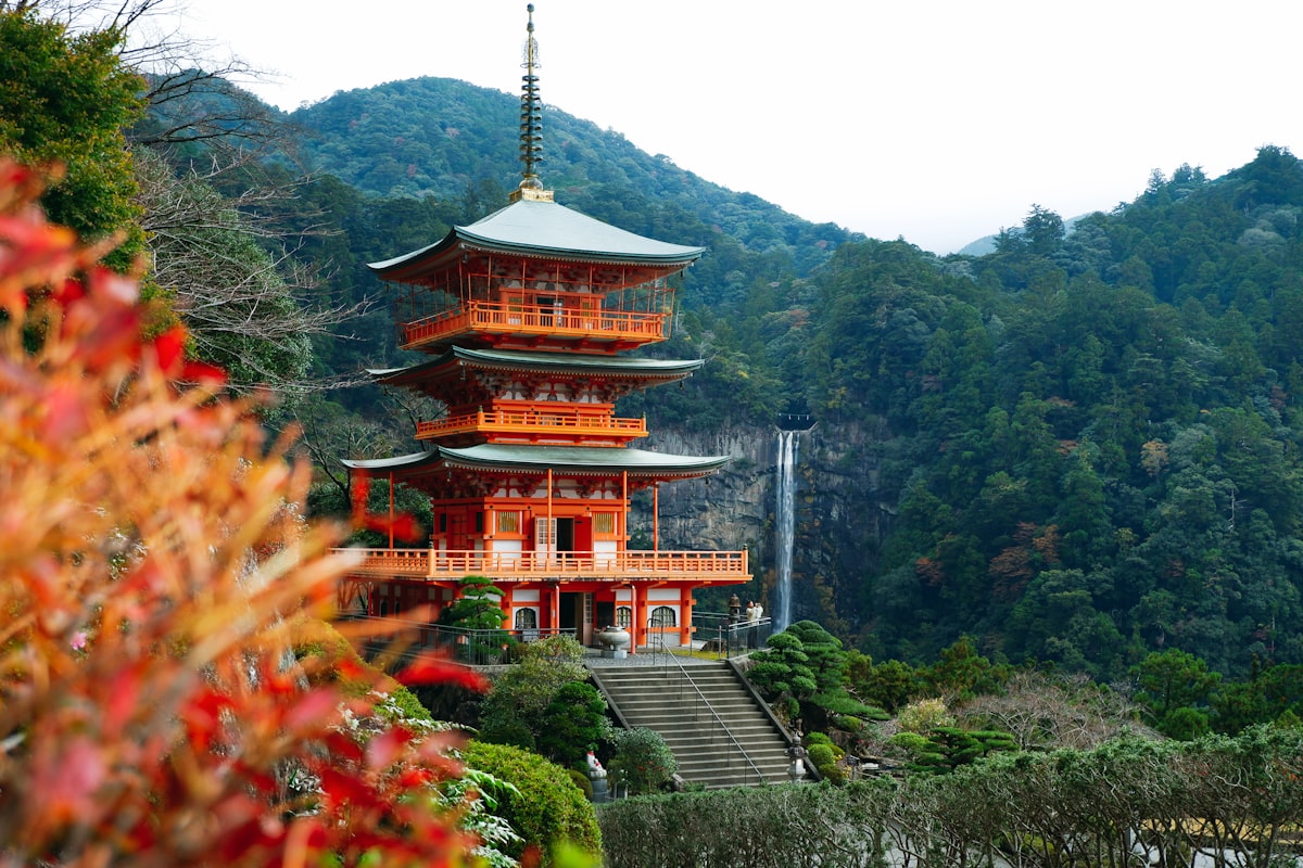Vermilion three-story pagoda framed by red autumn leaves with a tall waterfall dropping through forested hills behind