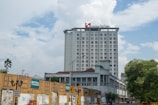 A tall hotel building with the name 'Swiss Belinn' on the top in a city environment. The forefront shows a construction wall with signs and graffiti, while the background features a partly cloudy sky.