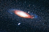 A spiral galaxy set against the vastness of space, featuring a bright, central core with arms that display a mix of reddish and bluish hues. Countless stars are scattered throughout the scene, contributing to a sense of infinite depth and wonder.
