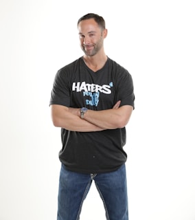A man with short hair and facial hair smiles confidently while crossing his arms. He wears a dark T-shirt with a slogan and blue jeans. The background is plain and bright.
