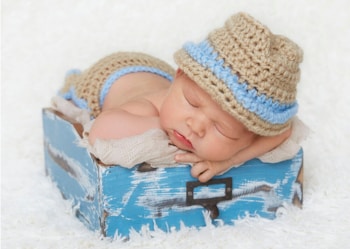A newborn baby is peacefully sleeping on its stomach, wearing a knitted beige and blue hat and matching diaper cover. The baby is resting on a soft blue and white textured prop that resembles a wooden box, with a fluffy white background enhancing the serene atmosphere.