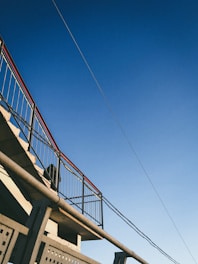 A metal staircase with railings is seen against a clear blue sky. The structure includes straight lines and geometric patterns, with a wire or cable extending across the top corner.