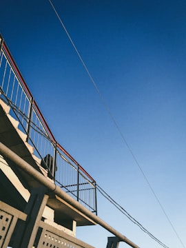 A metal staircase with railings is seen against a clear blue sky. The structure includes straight lines and geometric patterns, with a wire or cable extending across the top corner.