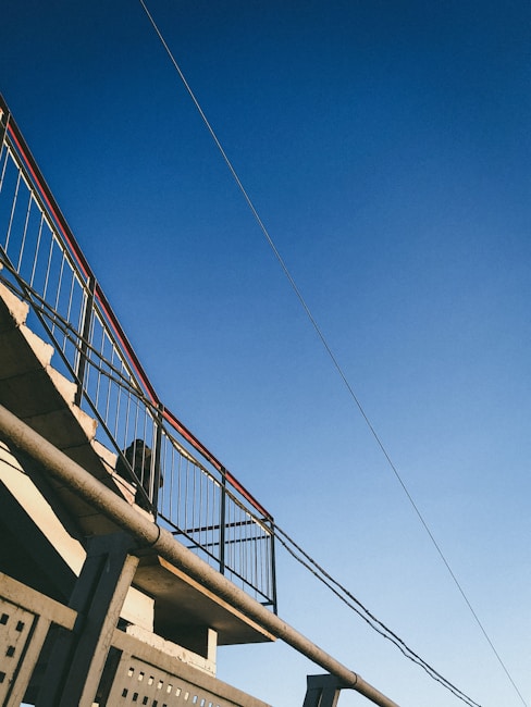 A metal staircase with railings is seen against a clear blue sky. The structure includes straight lines and geometric patterns, with a wire or cable extending across the top corner.