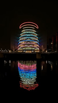 A modern architectural structure illuminated with vibrant LED lights in a variety of colors, including red, blue, and green. The building is reflected in the calm water of a river or canal below, creating a symmetrical and colorful reflection. The scene is set against a dark night sky, emphasizing the brightness and vividness of the lights.