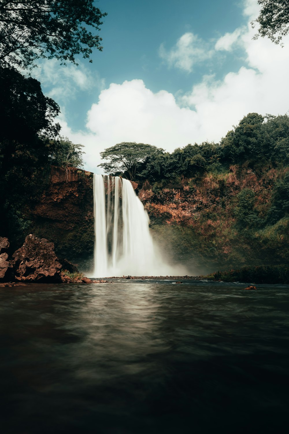 waterfalls near green trees under white clouds and blue sky during daytime