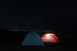 Two tents are set up under a starry night sky, with one glowing from an interior light, casting an orange hue. The background reveals distant city lights on the horizon, creating a serene camping atmosphere.