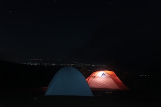 Two tents are set up under a starry night sky, with one glowing from an interior light, casting an orange hue. The background reveals distant city lights on the horizon, creating a serene camping atmosphere.