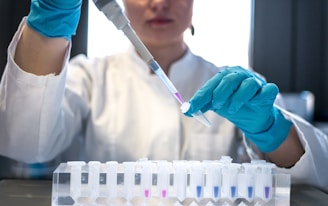 A lab technician wearing a white coat and blue gloves is carefully transferring a liquid with a pipette into a small test tube. Several other test tubes are organized in a rack in the foreground, some containing various levels of liquid.