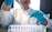 A lab technician wearing a white coat and blue gloves is carefully transferring a liquid with a pipette into a small test tube. Several other test tubes are organized in a rack in the foreground, some containing various levels of liquid.