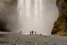 A group of people stands near a large, roaring waterfall surrounded by moss-covered cliffs. The mist from the waterfall creates an ethereal atmosphere. The ground is rocky and wet, indicating proximity to the water.