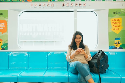 A woman is sitting comfortably on a bright blue seat inside a train, focusing on her smartphone with a slight smile. She is casually dressed, wearing a beige sweater and jeans, with a backpack placed beside her. The train interior is decorated with colorful signs and posters.