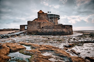 A large, stone fortress with a round tower stands surrounded by rocky terrain and scattered pools of water. The fortress appears old and weathered, with scaffolding on part of the structure, suggesting ongoing restoration work. The sky is filled with clouds, casting a muted light over the scene.