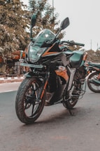 A sleek, black and orange sports motorcycle is parked on a road. It has a prominent front fairing and visible brand decals. The bike's design is aerodynamic and modern, with large tires and mirrors extending outward. In the background, there is some greenery and another motorcycle partially visible.