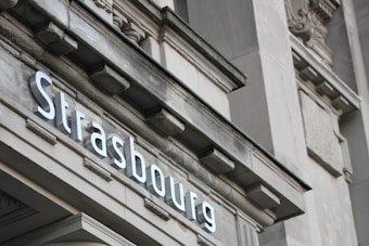 A close-up view of an architectural structure with the word 'Strasbourg' displayed in bold, white letters. The building features decorative elements and a classic design, with stone details and a slightly weathered texture.
