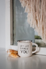 A white ceramic mug with the words 'LET LIFE SURPRISE YOU' printed on it is placed on a smooth surface. Next to it, two seashells are arranged. Behind the mug, there is a mirror partially covered by a light-colored fringe or macrame decoration.