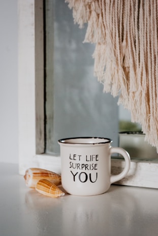 A white ceramic mug with the words 'LET LIFE SURPRISE YOU' printed on it is placed on a smooth surface. Next to it, two seashells are arranged. Behind the mug, there is a mirror partially covered by a light-colored fringe or macrame decoration.
