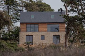 A modern two-story house with a combination of stone and wood paneling. Large windows dominate the upper floor, allowing ample natural light. The roof is covered with dark shingles, and the property is surrounded by tall trees which add to the serene ambiance. The house sits in a natural setting with some sparse shrubbery in the foreground.