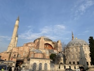 A historic architectural structure featuring a large dome and several minarets under a clear blue sky. The building exterior has a mix of stone and brickwork, with arched windows and intricate details.
