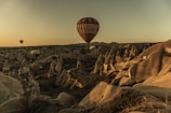 Hot air balloons soar over a unique rocky landscape at sunset, with warm hues casting dramatic shadows on the terrain below. The scene appears serene as the balloons float in the quiet, expansive sky.