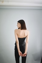 A person standing with their back to the camera, wearing a black corset and black trousers. The background is a plain, light grey wall, creating a minimalist and elegant look.