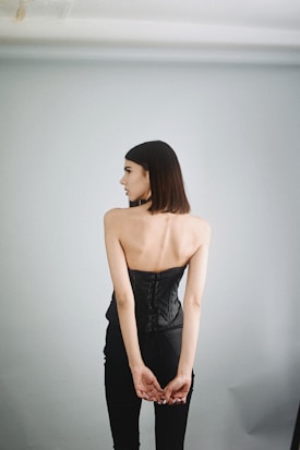 A person standing with their back to the camera, wearing a black corset and black trousers. The background is a plain, light grey wall, creating a minimalist and elegant look.