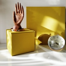 A wooden hand sculpture is placed on top of a yellow box, accompanied by a colorful bracelet at the base of the sculpture. Next to it, there is an Omega clock with Roman numerals, leaning against a yellow backdrop. The scene is well-lit, with shadows and light reflections adding depth to the composition.