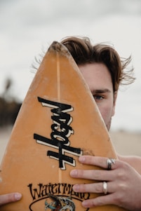 A person partially concealed by the top of a surfboard, with one eye visible. The surfboard features a prominent brand logo and a detailed graphic at the bottom. The individual is wearing multiple rings and has wind-swept hair.