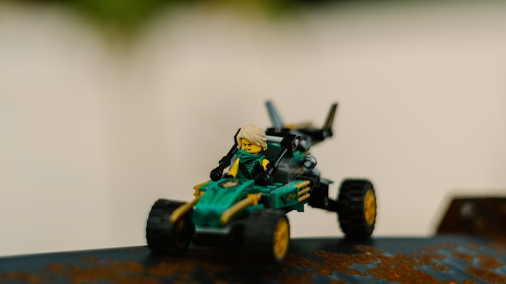 A small toy figure with blonde hair sits in a detailed, green and yellow toy vehicle with large black wheels. The vehicle appears to be a camouflaged combat or racing car, sporting intricate designs and attachments. The background is predominantly blurred, focusing on the toy as the main subject.