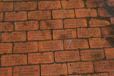 A series of engraved red bricks with various personal messages and dedications. The inscriptions often include references to the Boston Red Sox and expressions of loyalty and fandom, as well as dedications to family and friends.