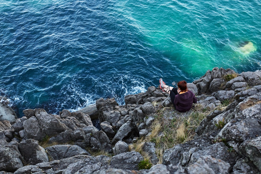 woman in black jacket and black pants sitting on rock by the sea during daytime